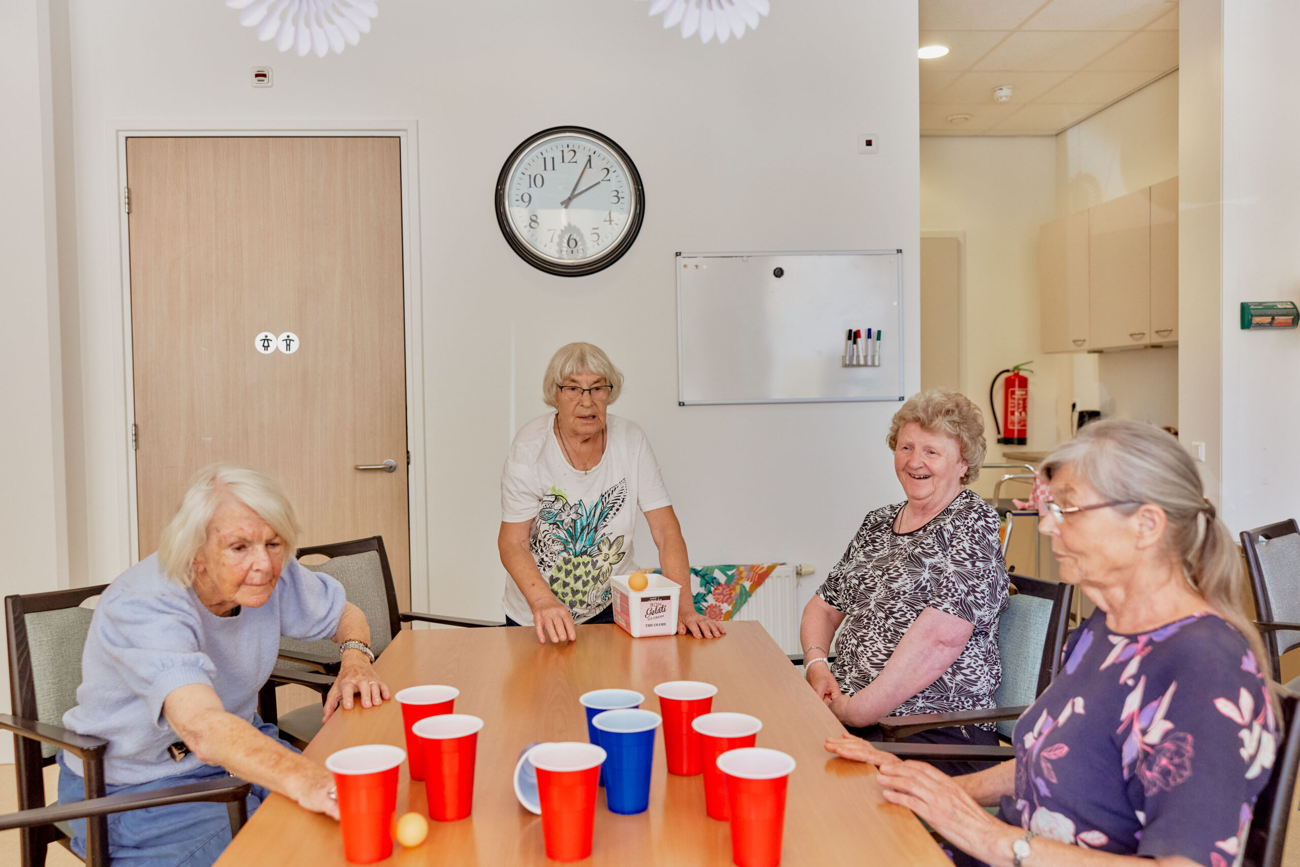 Huiskamer/keuken in woon-zorgcentrum. Drie oudere vrouwen zitten rondom een tafel waar rode en blauwe bekertjes op staan. Eén vrouw gooit een pingpongballetje richting de bekers.