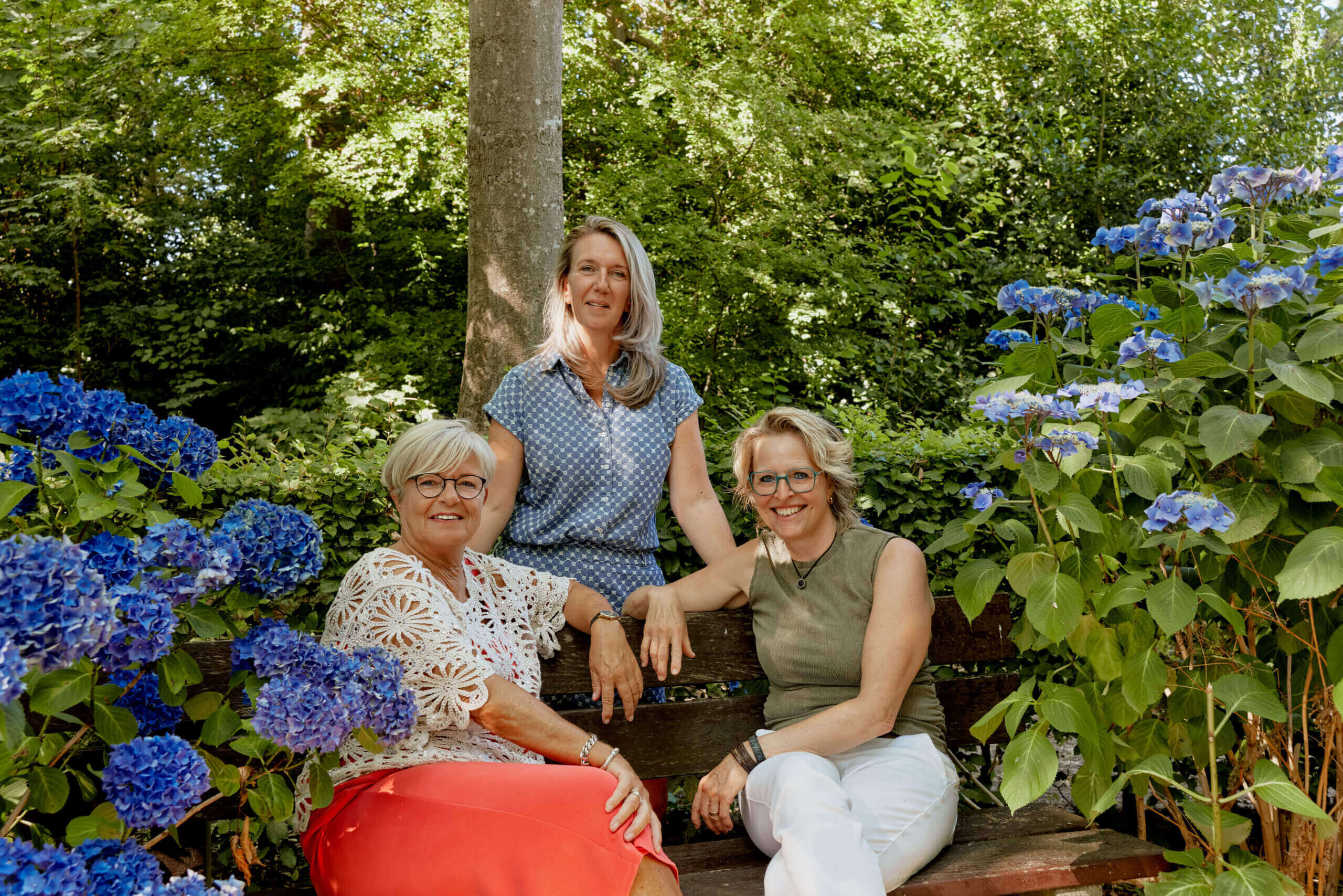Foto van 3 betrokkenen RAP-ZHN, tussen groene bomen en struiken en blauwe bloemen. Lachend in de camera.